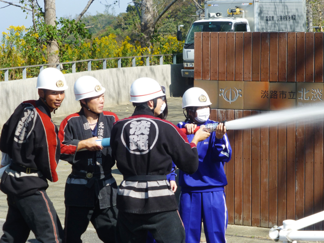 消防団のサポートを受けて、校門前から斜面に向かって放水訓練を行う女子生徒。
